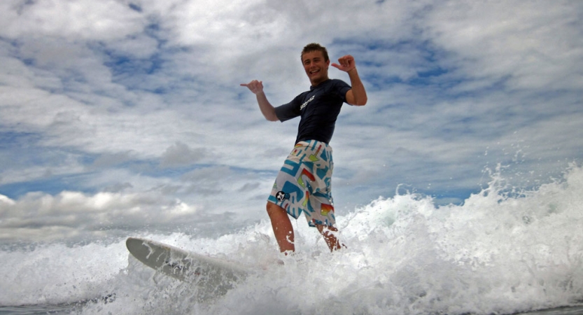 a person surfs on a white wave, smiles and gives the camera two "hang loose" signs